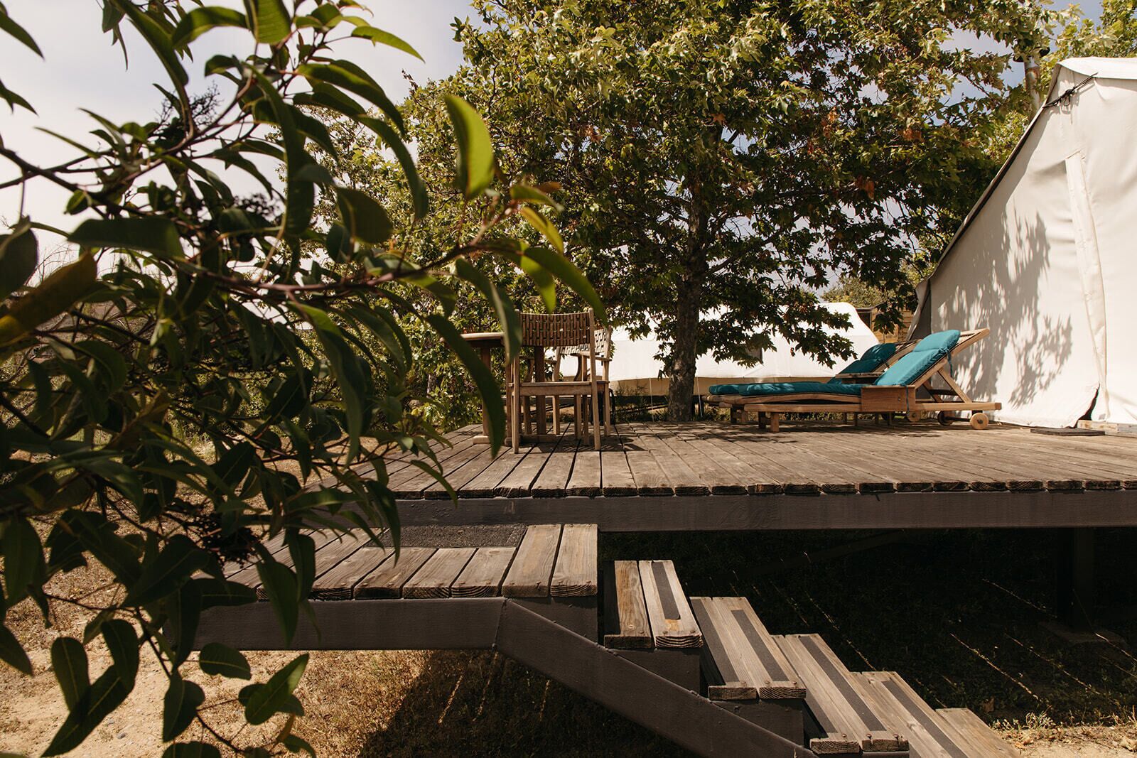 A wooden deck with a chair and a lounge chair, a tree, and a tent.