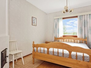 A bedroom with a wooden bed, a chair, a small bedside table, a window with curtains, and a wall-mounted picture.