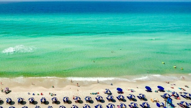 A beach with clear turquoise water and a sandy shore. There are multiple beach umbrellas and lounge chairs arranged along the beach.