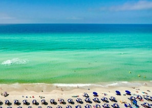 A beach with clear turquoise water and a sandy shore. There are multiple beach umbrellas and lounge chairs arranged along the beach.