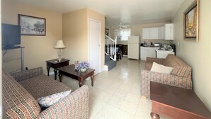 A living room with patterned sofas, a dining table, a TV, and a kitchen in the background.