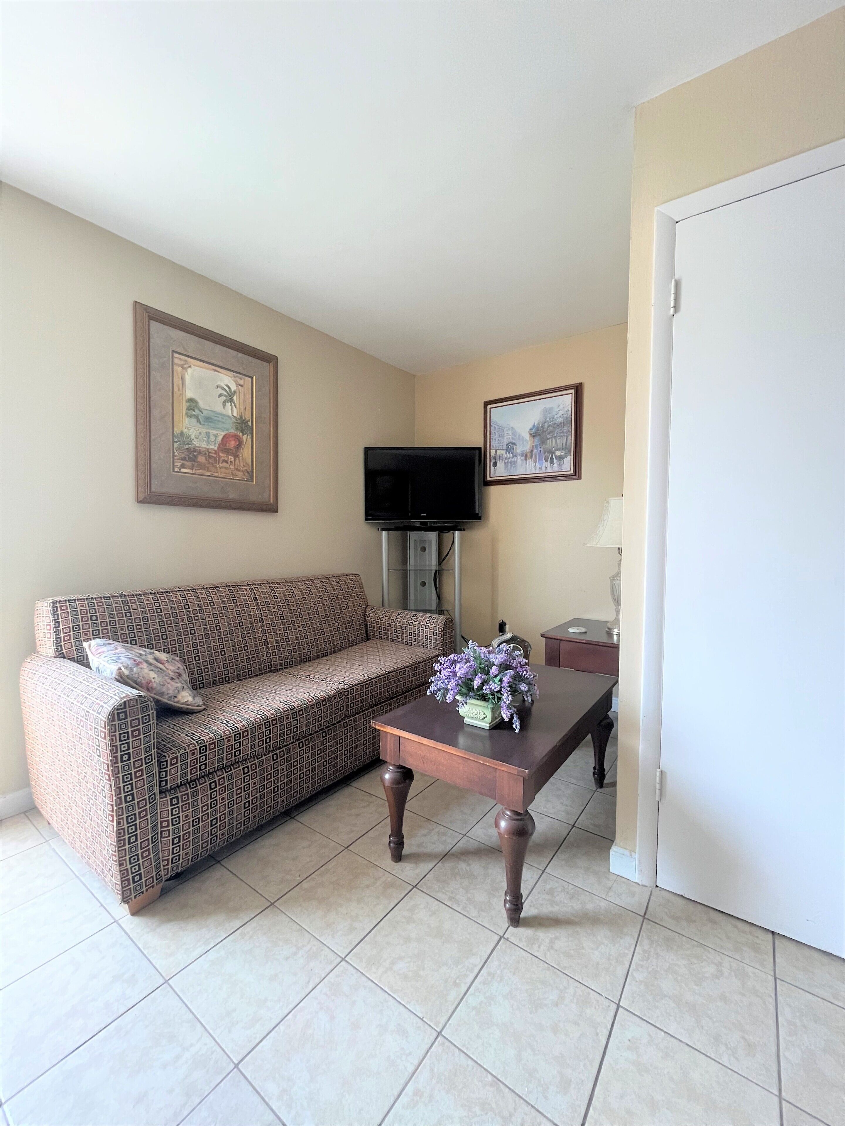 A living room with a patterned sofa, a coffee table with a vase of flowers, a flat-screen TV mounted on the wall, and a framed picture above the TV.