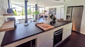 Une cuisine moderne dotée d’un grand îlot central, d’appareils électroménagers intégrés et de grandes fenêtres offrant une vue sur l’extérieur.