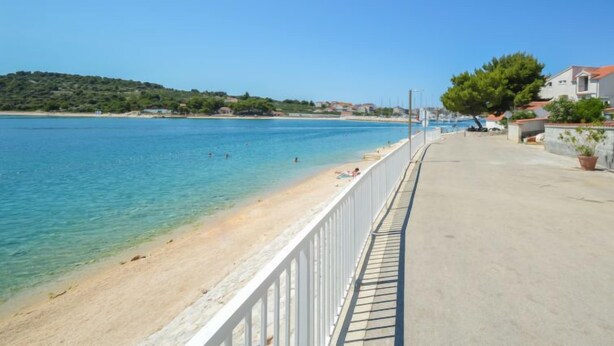 A coastal walkway with a clear view of the sea, a sandy beach, and residential buildings.