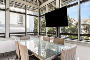 Una habitación iluminada por el sol, con una mesa de vidrio, sillas de mimbre, ventanas amplias y un televisor de pantalla plana montado en la pared.