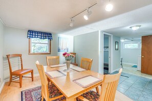 Een eetruimte met een houten tafel, stoelen, een vaas met bloemen en een raam met een blauw-witte geruit gordijn.