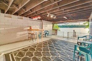 Een overdekt terras met houten plafond, bakstenen muur en uitzicht op een meer. Er staan stoelen en een tafel met een plant erop.