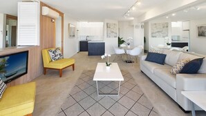 Una sala de estar moderna con un sofá, un sillón y una mesa de centro. Hay un televisor a la izquierda y una cocina al fondo.