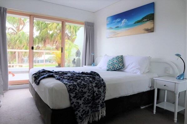 A bedroom with a bed, a nightstand, a painting, and a view of palm trees and a beach.