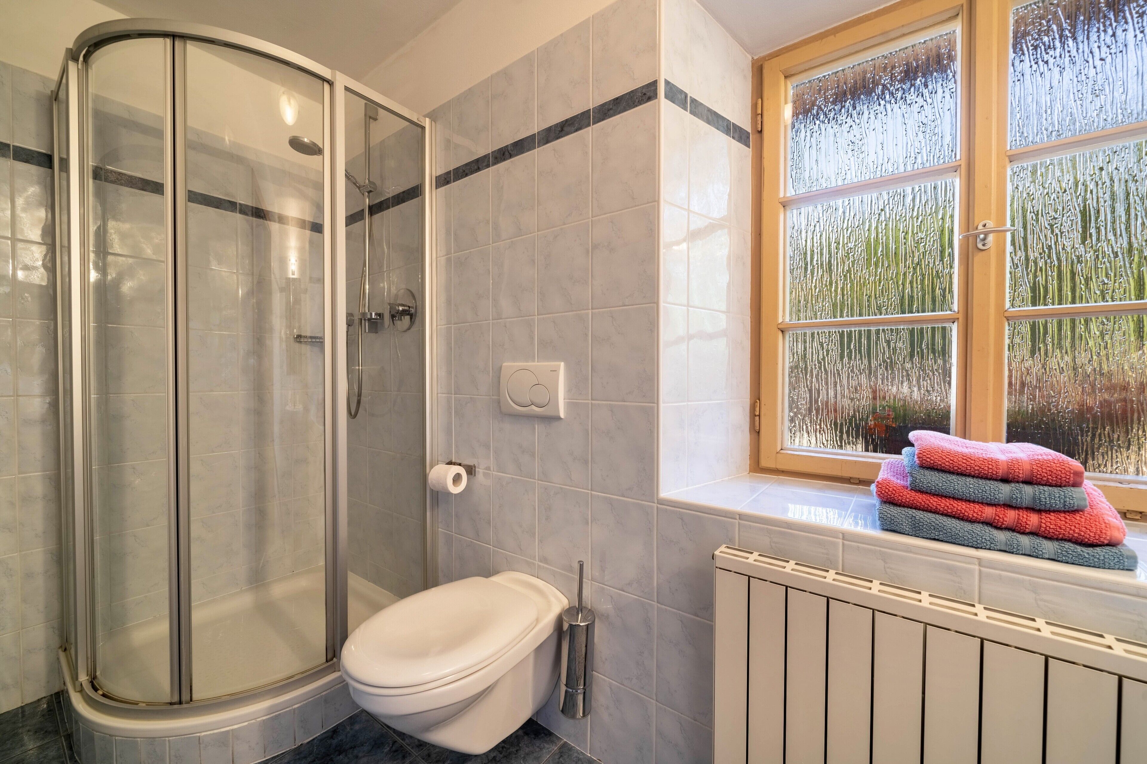 Et badeværelse med bruseniche, toilet, et vindue med udsigt og en radiator.