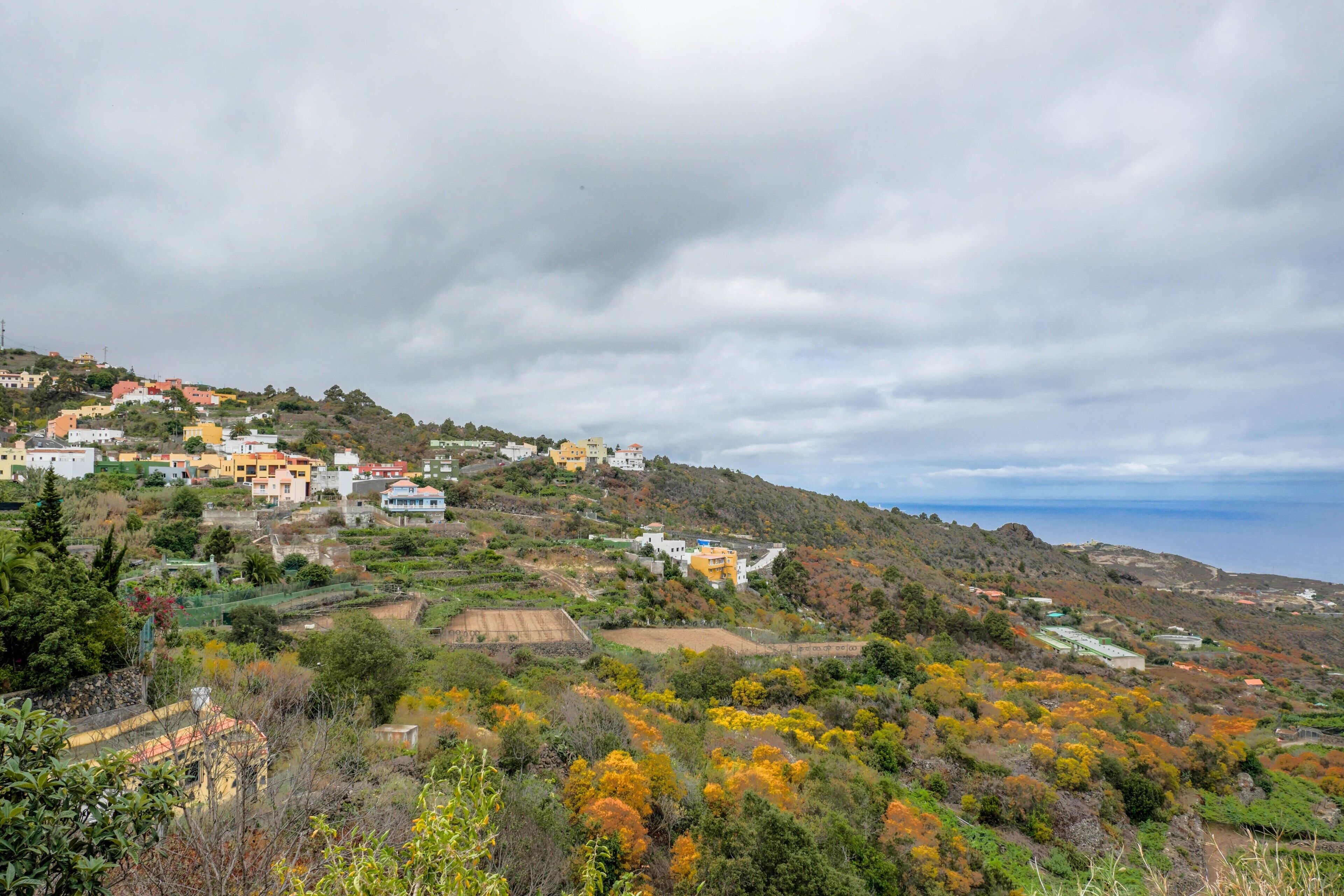 A coastal village with buildings on a hillside, a view of the sea, and autumn-colored trees.