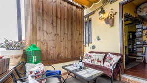 A cozy patio area with a wooden fence, a cushioned sofa, a small table with a bottle and glasses, and a green crate.