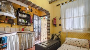 A compact room with a bed, a bench, a small table, a shelf with dishes, a fire extinguisher, and a window with curtains.