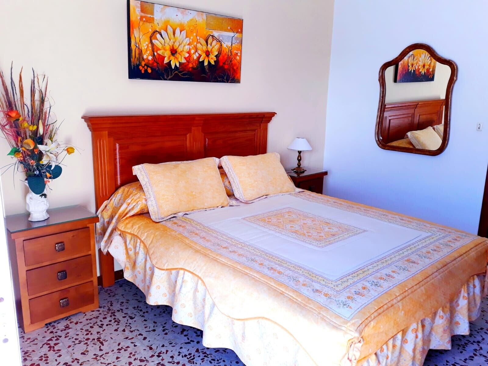 Een slaapkamer met een bed, nachtkastje, lamp, spiegel en een schilderij aan de muur.