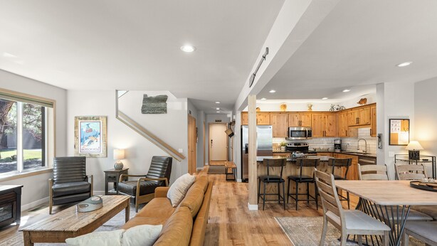 A spacious living area with a sofa, chairs, a coffee table, and a dining table with chairs. A kitchen with wooden cabinets and appliances is visible in the background. - Newly Remodeled Townhouse, 2 Master Suites, Hot Tub, SHARC Passes, Bikes (Sunriver)