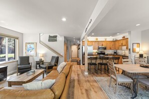 A spacious living area with a sofa, chairs, a coffee table, and a dining table with chairs. A kitchen with wooden cabinets and appliances is visible in the background. - Newly Remodeled Townhouse, 2 Master Suites, Hot Tub, SHARC Passes, Bikes (Sunriver)