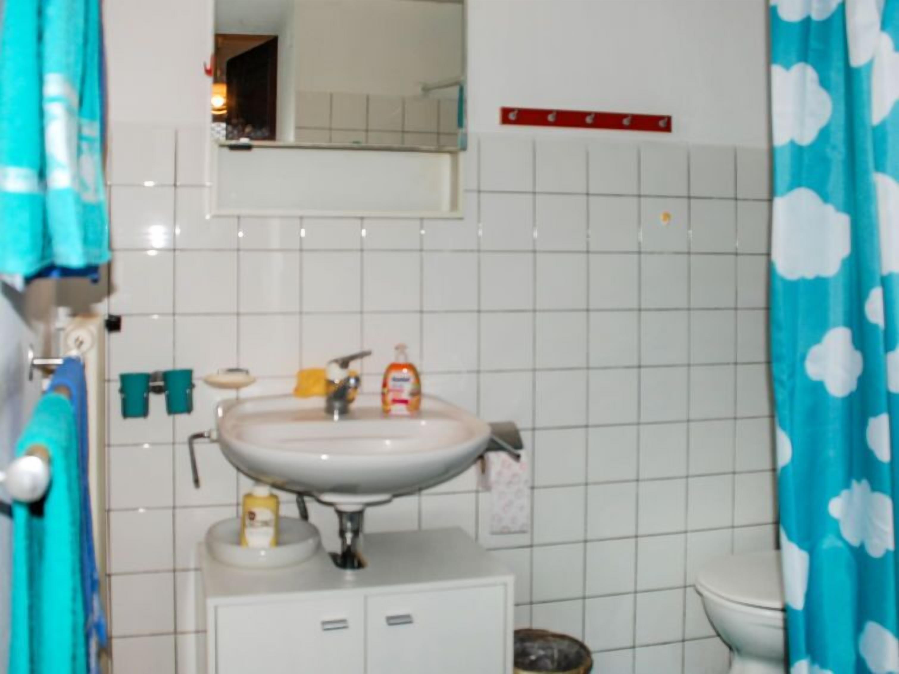Casa de banho com lavatório branco, espelho e sanita. Há artigos de higiene pessoal no lavatório e uma toalha pendurada na parede.