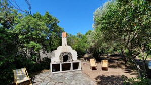 A stone patio with a stone oven, two wooden lounge chairs, and surrounding greenery.