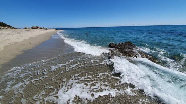 A beach with clear blue water, a sandy shore, and rocks near the water's edge.
