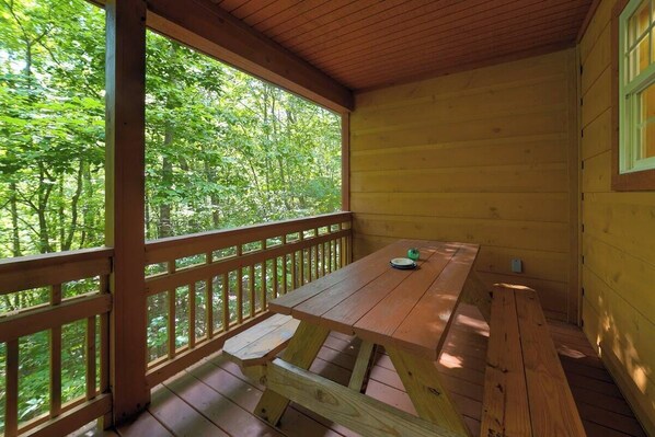 A wooden deck with a picnic table and benches, surrounded by a wooden railing, overlooking a forested area.