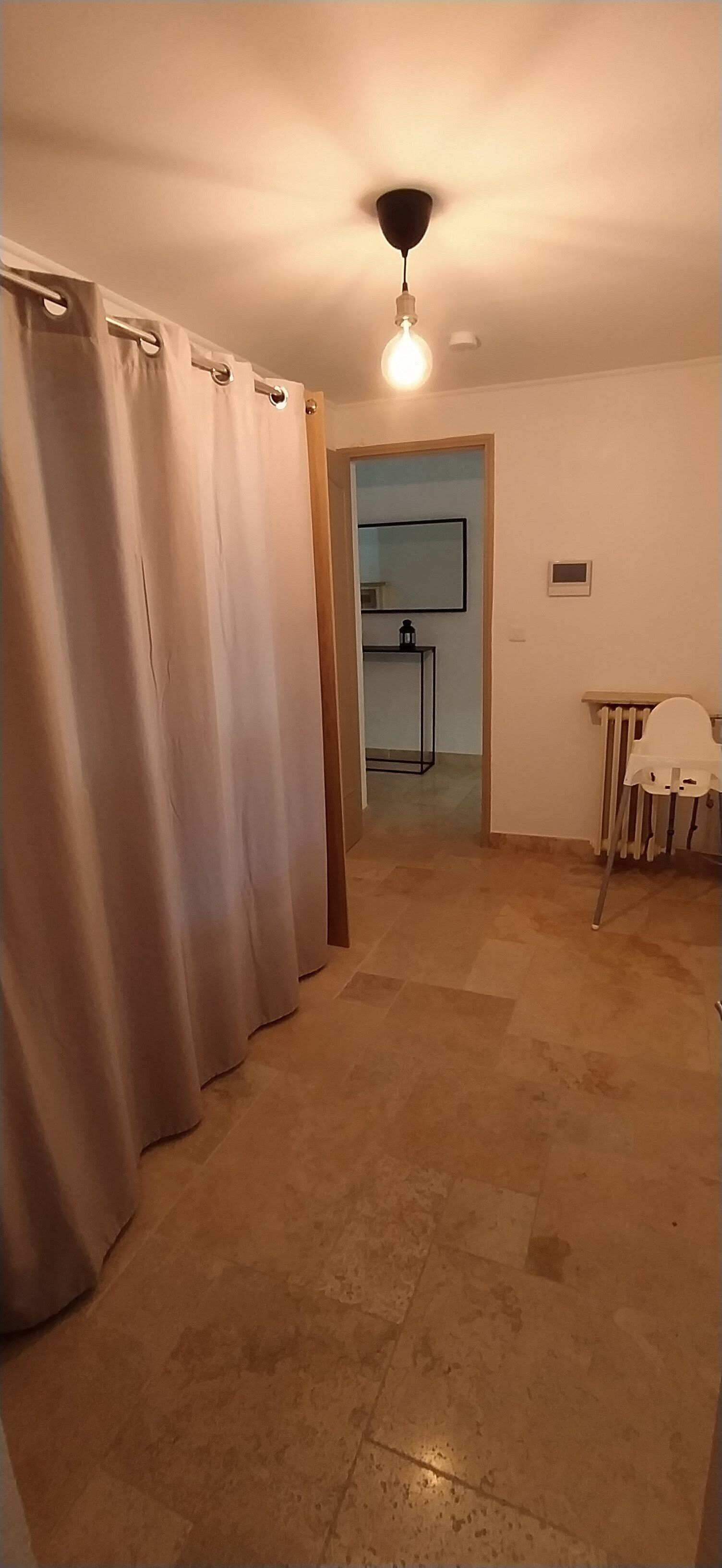 A room with a tiled floor, a ceiling fan, a radiator, and a door leading to a kitchen area.