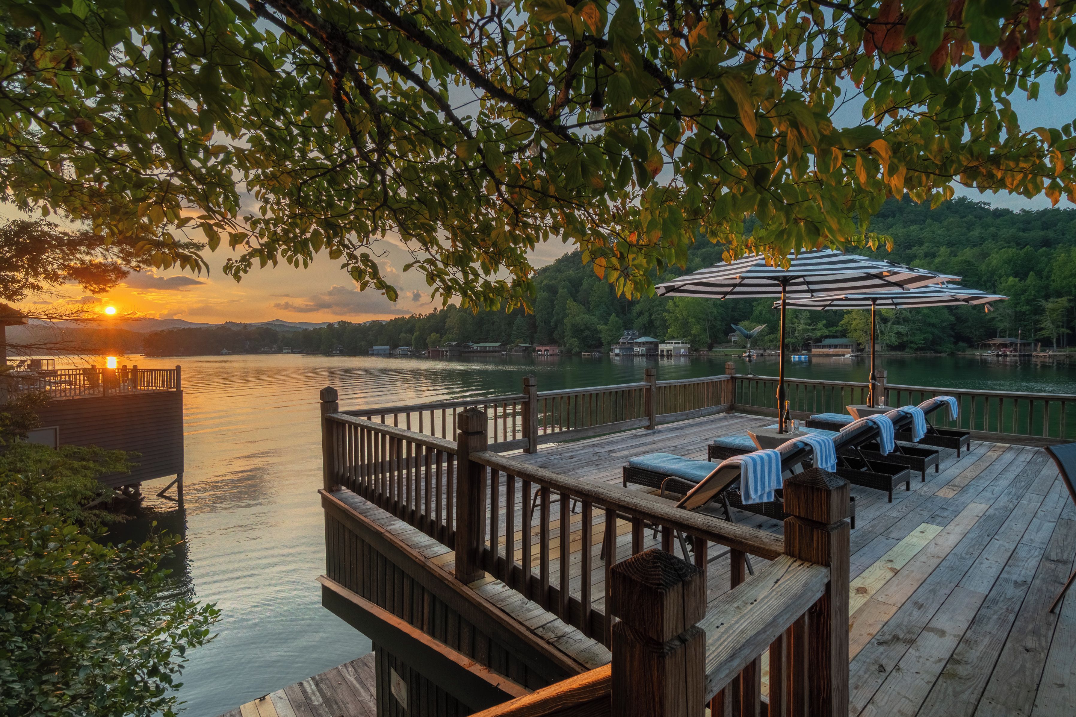 A wooden deck with lounge chairs and an umbrella, overlooking a calm body of water with a sunset in the background.