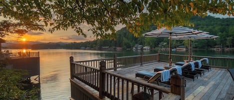 A wooden deck with lounge chairs and an umbrella, overlooking a calm body of water with a sunset in the background.