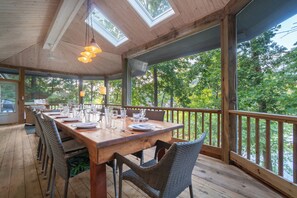 Eine Holzterrasse mit einem langen Esstisch, umgeben von Korbstühlen, mit Blick auf ein bewaldetes Gebiet.