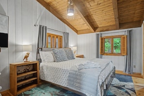 Une chambre avec un plafond en bois, un lit recouvert d’une couvre-lit à motifs, deux tables de chevet avec des lampes et une fenêtre avec des volets.