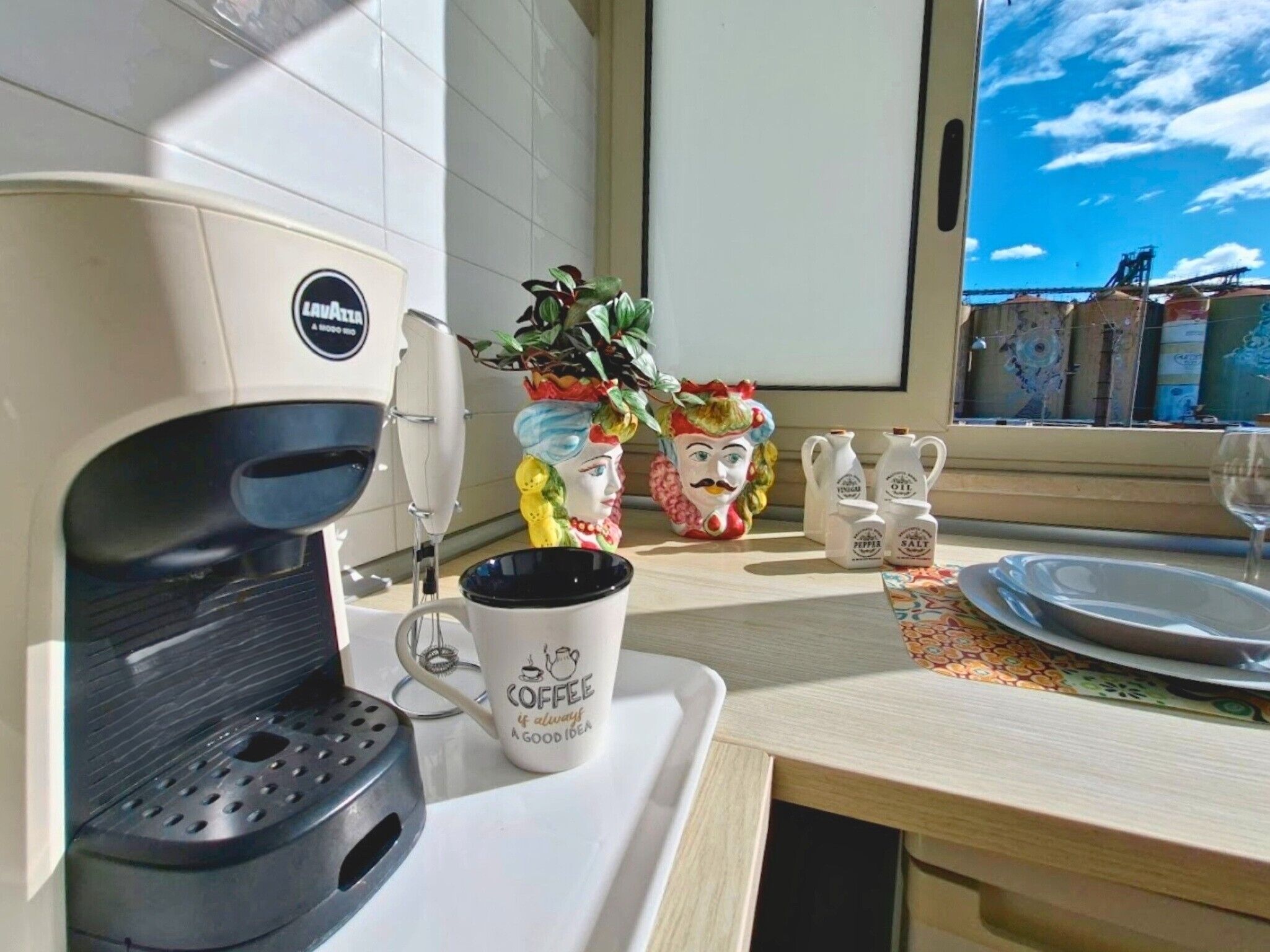 Una cocina con cafetera, una planta, saleros y pimenteros, y un ventanal con vistas al cielo y edificios.