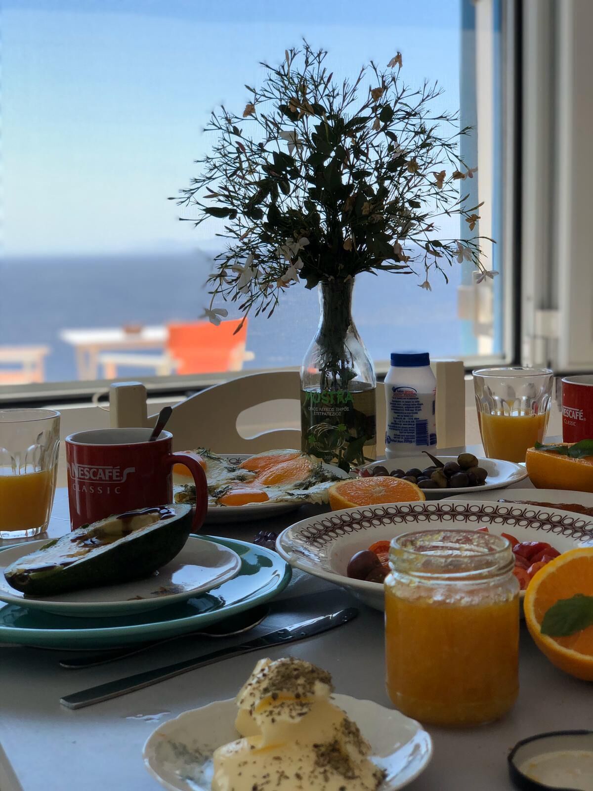 A breakfast table with a vase of flowers, plates of food, and a view of the sea.