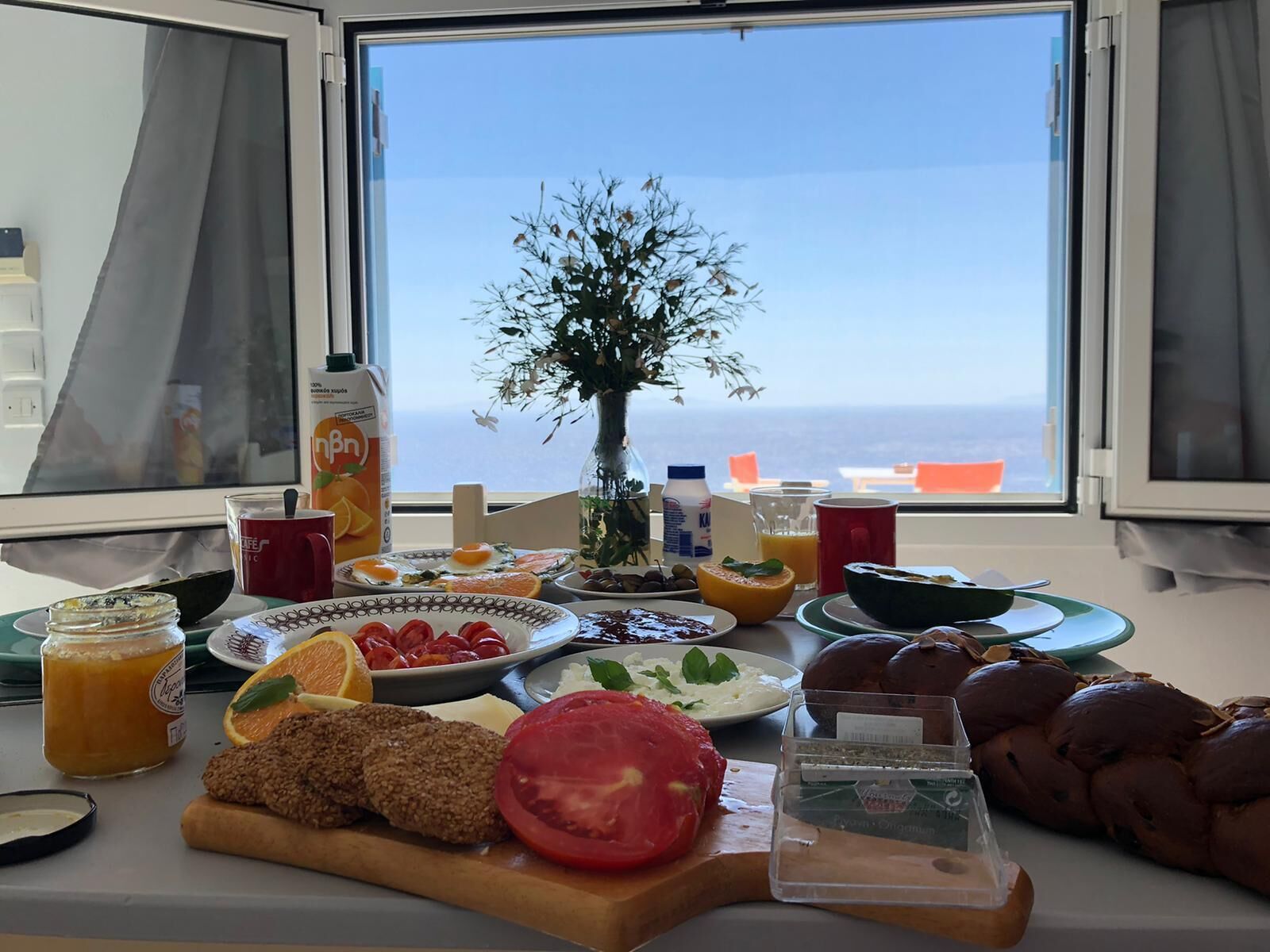 A breakfast table with various foods and drinks, a vase with flowers, and a view of the sea through a window. - Quiet private house with Sea view    (Sifnos)