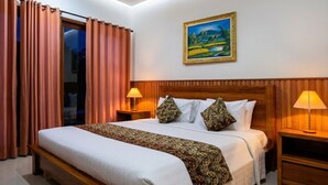 A hotel room with a large bed, two bedside lamps, a painting on the wall, and a view through a window with curtains. - Cening Ayu Sanur (Denpasar)