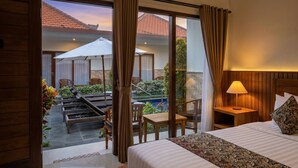 A hotel room with a bed, a nightstand, a lamp, a view of an outdoor patio with a pool, and a dining area. - Cening Ayu Sanur (Denpasar)