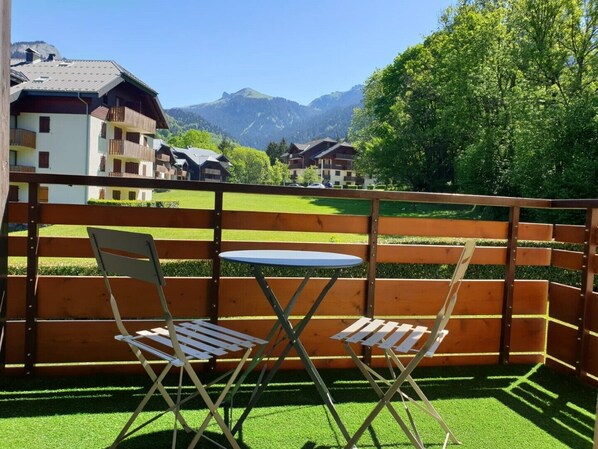A balcony with two wooden chairs and a small round table, overlooking a green lawn and a mountain range.