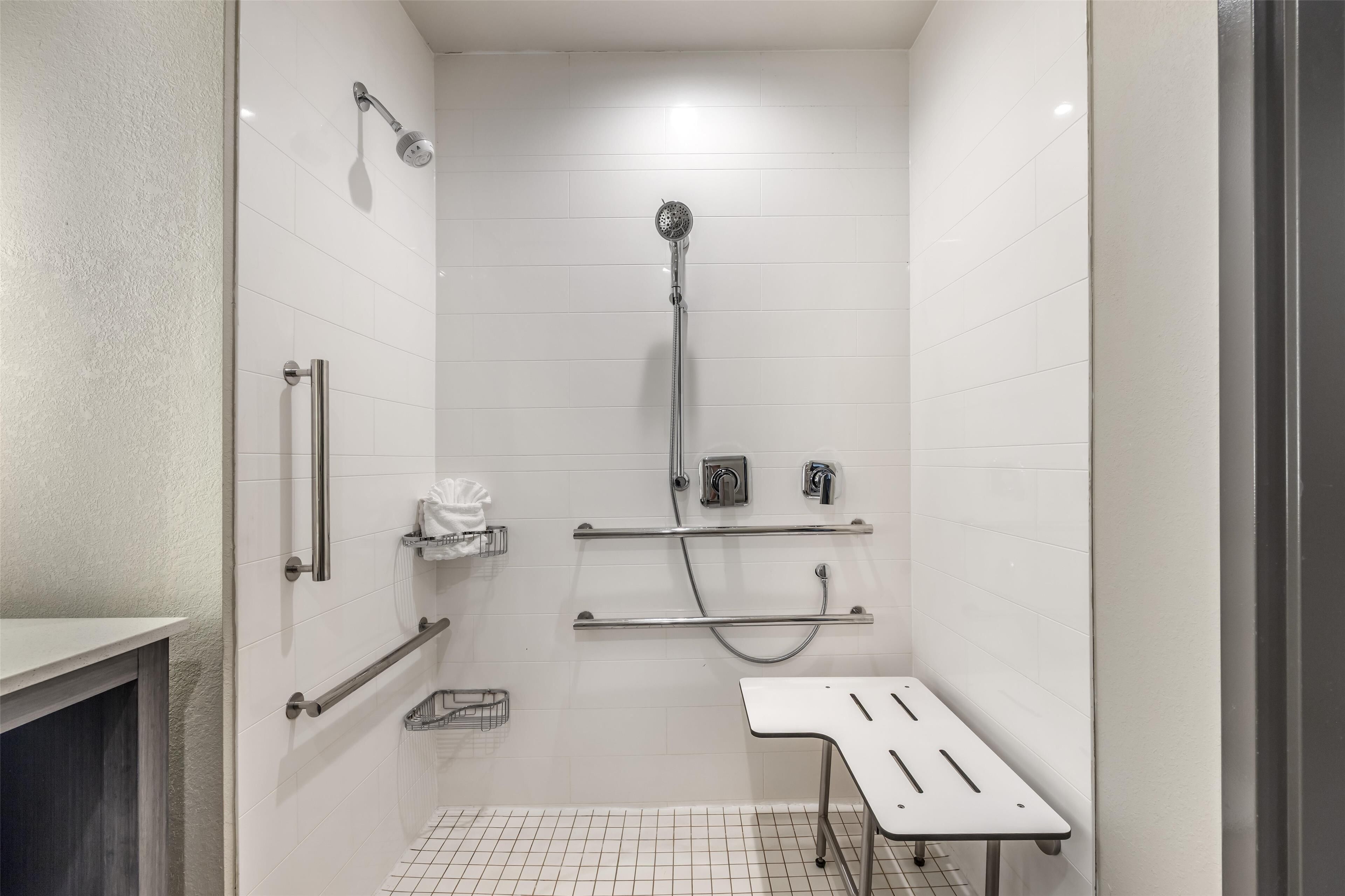 Un baño moderno con paredes de azulejos blancos, ducha con cabezal de ducha manual, barras de apoyo, toallero y un banco.