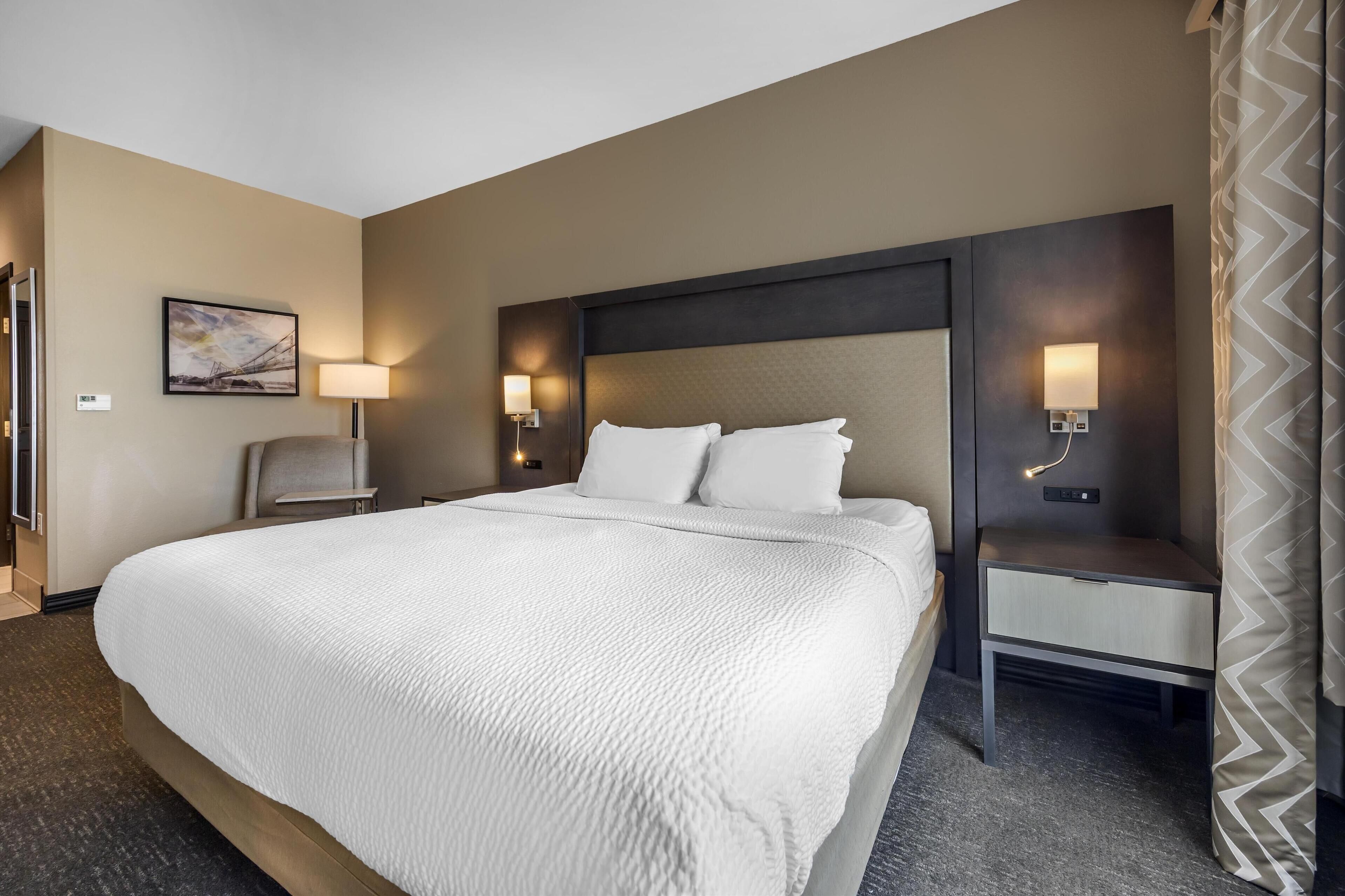 Ein Hotelzimmer mit einem großen Bett, einem Nachttisch mit Lampe, einer Wandleuchte und einem Gemälde.