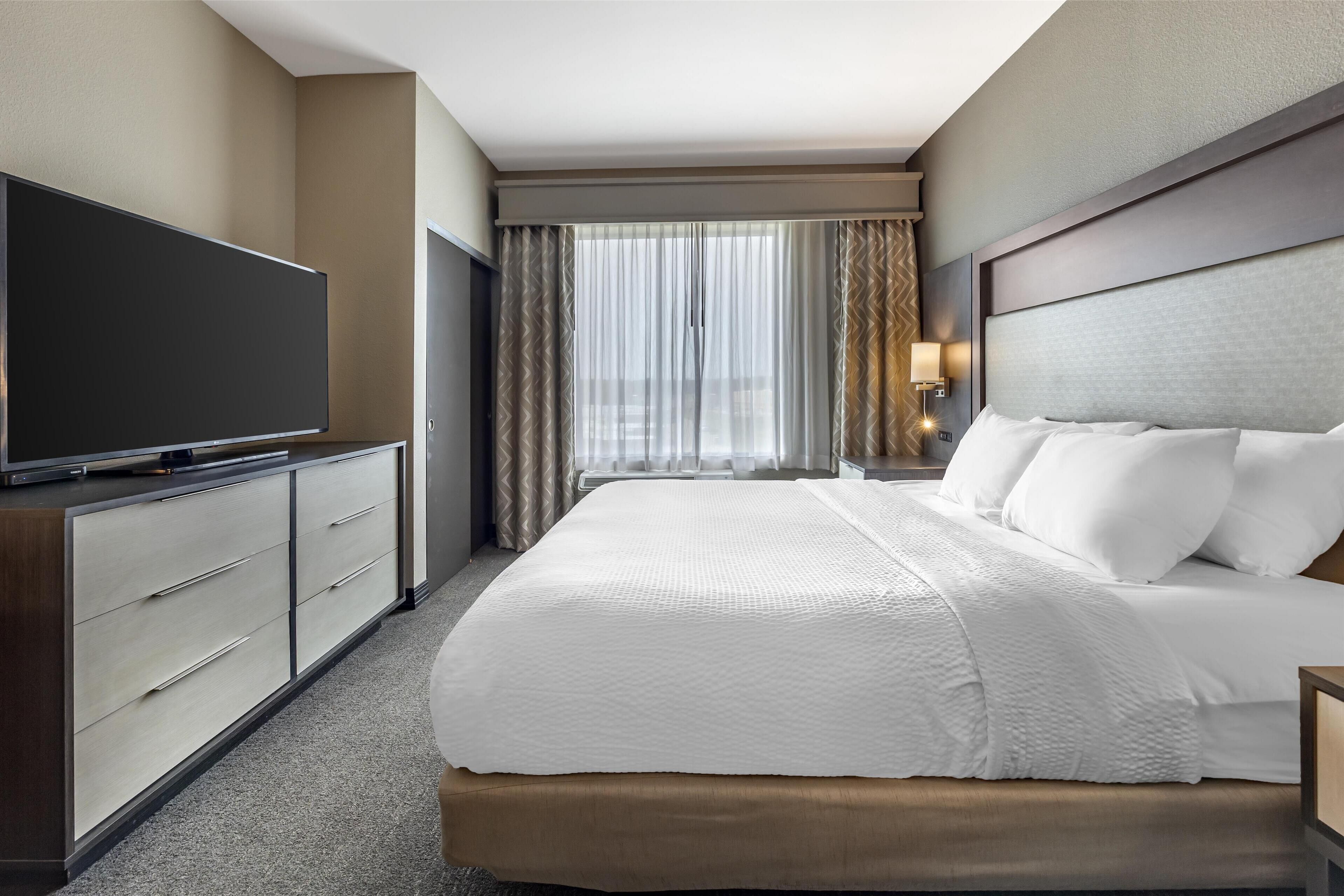 A hotel room with a large bed, a flat-screen TV, a desk with a lamp, and a window with sheer curtains.