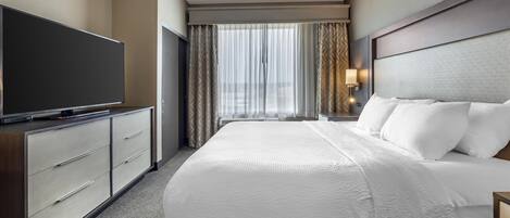 A hotel room with a large bed, a flat-screen TV, a desk with a lamp, and a window with sheer curtains.