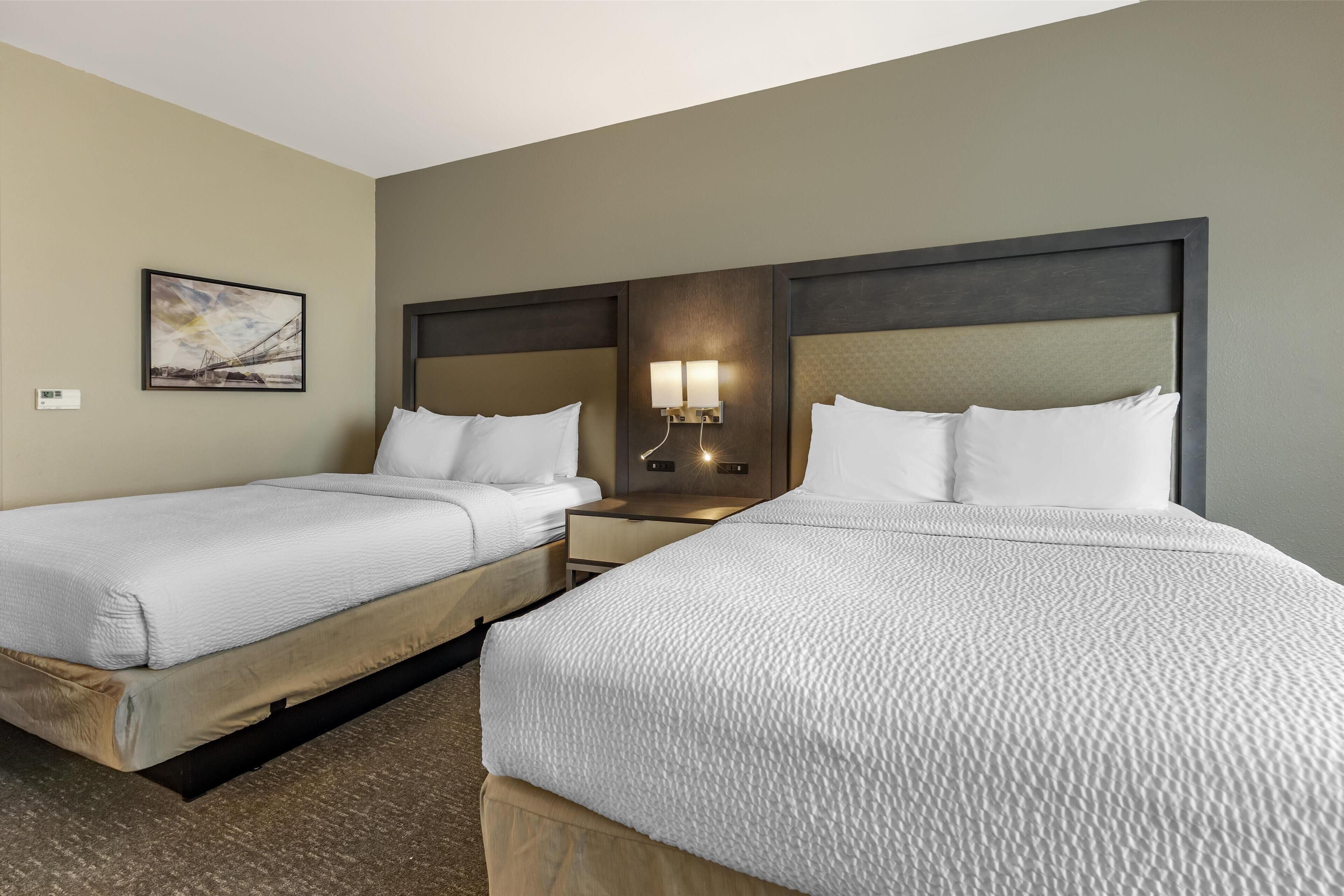 A hotel room with two beds, a nightstand, a wall-mounted lamp, and a framed picture on the wall.