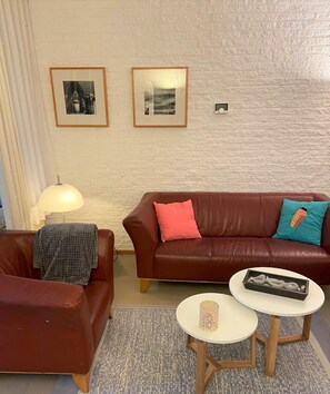 A living room with a brown leather sofa, a matching armchair, a small white table, and two framed pictures on the wall.