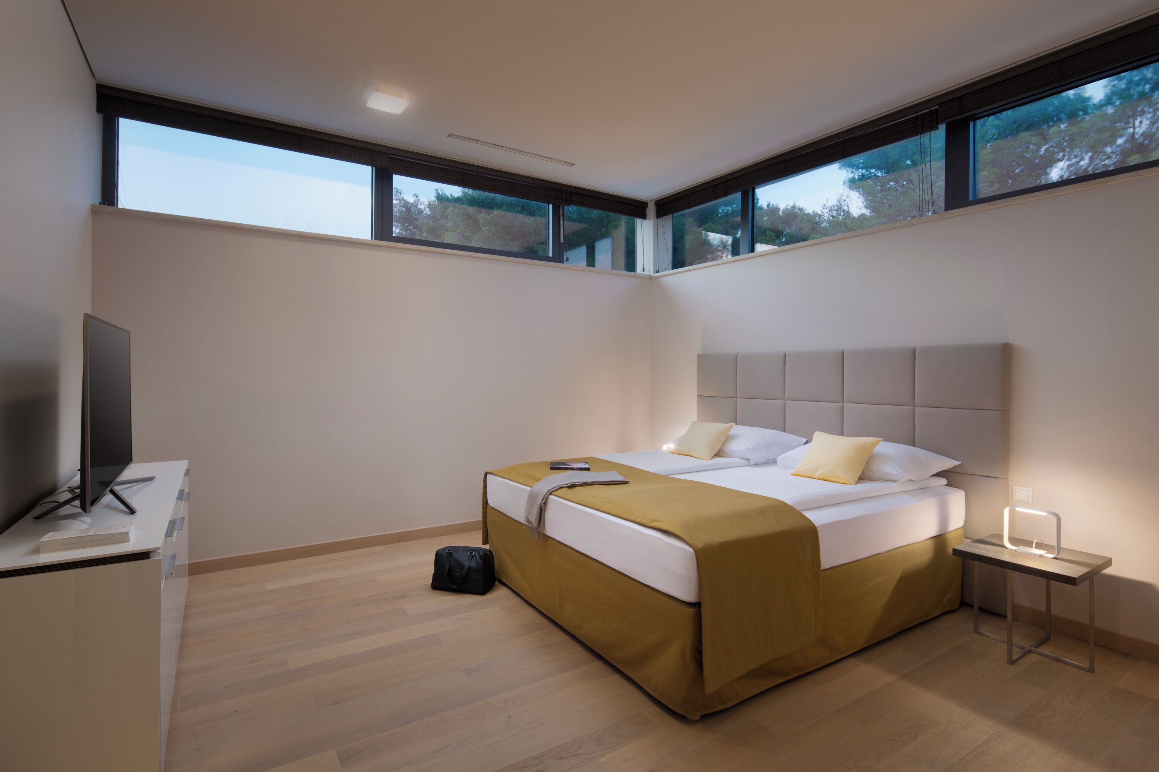 A modern hotel room with a bed, a desk with a TV, and large windows.