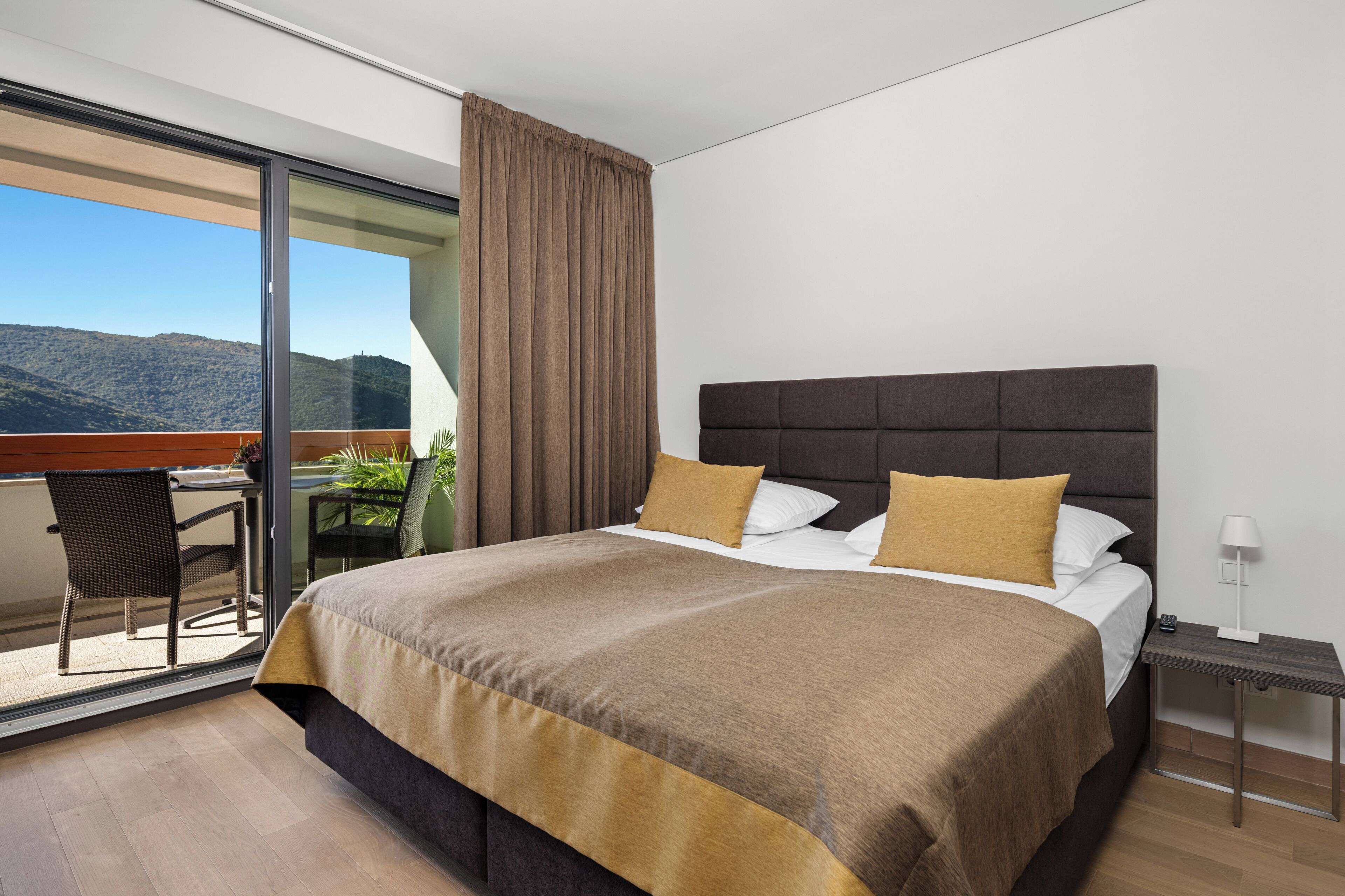 A modern hotel room with a large bed, a small desk, and a balcony with a view of the mountains.