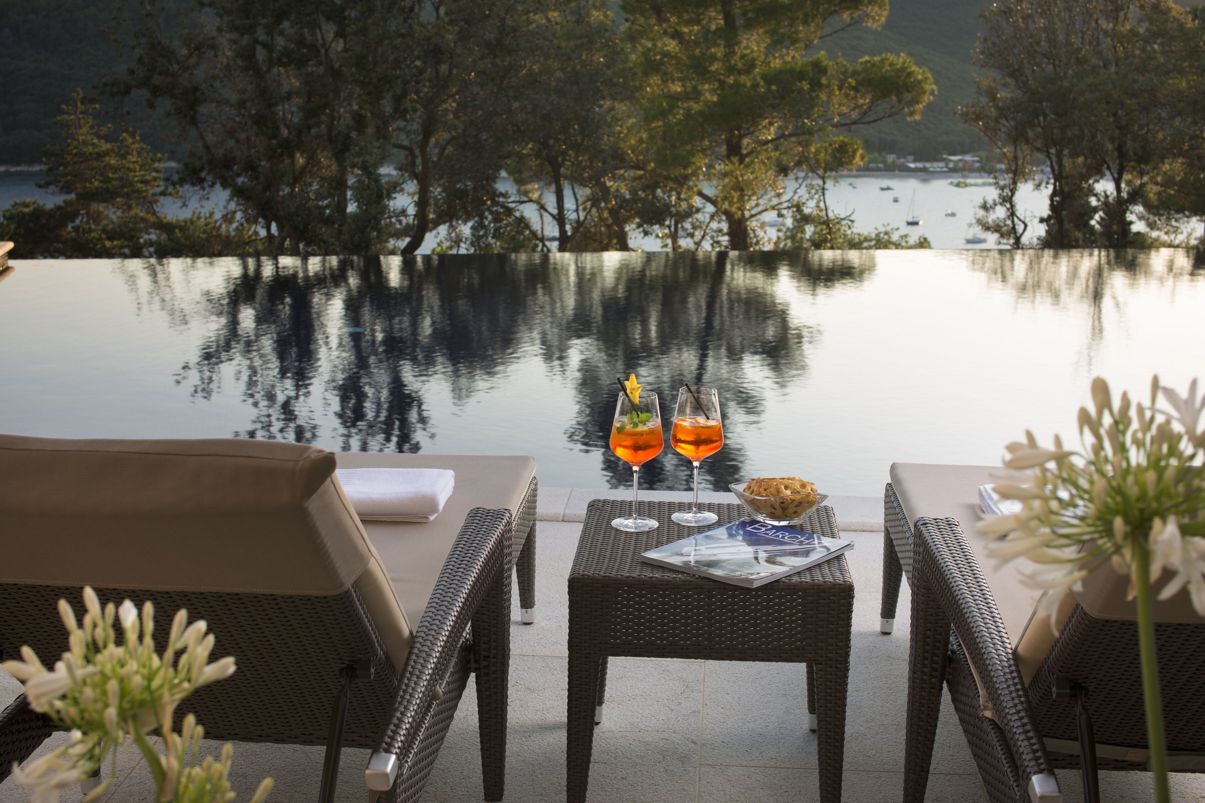 A serene outdoor seating area with wicker furniture and a table set with drinks and a menu, overlooking a calm body of water with boats.