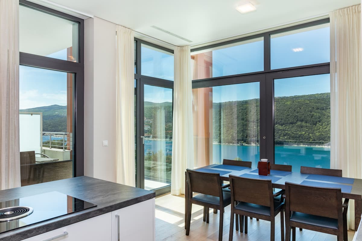 A modern kitchen with a large island, dining area with chairs, and large windows offering a view of a lake and mountains.
