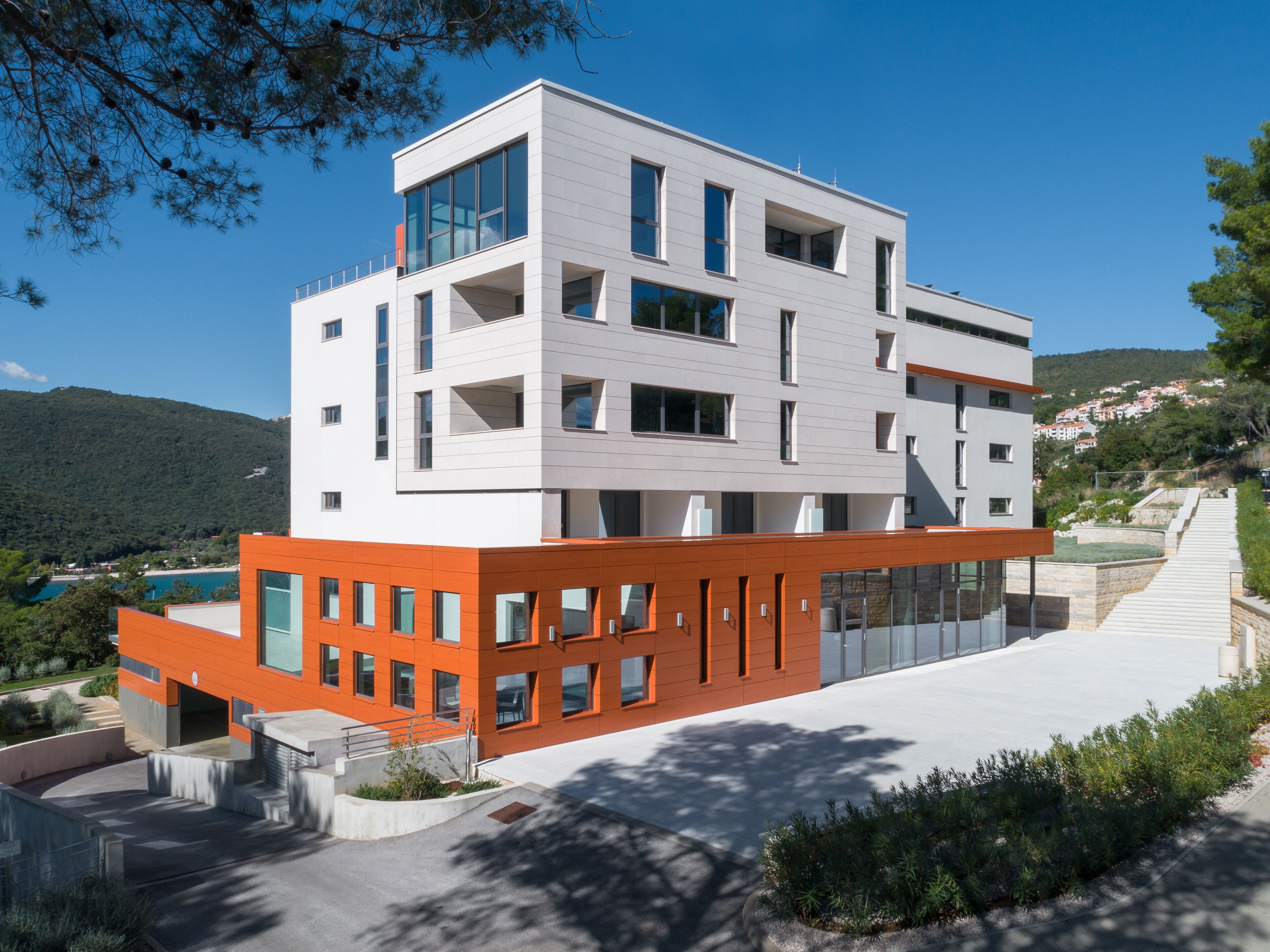 Un edificio moderno de varios pisos con techo plano, grandes ventanales y una combinación de paredes exteriores blancas y anaranjadas.