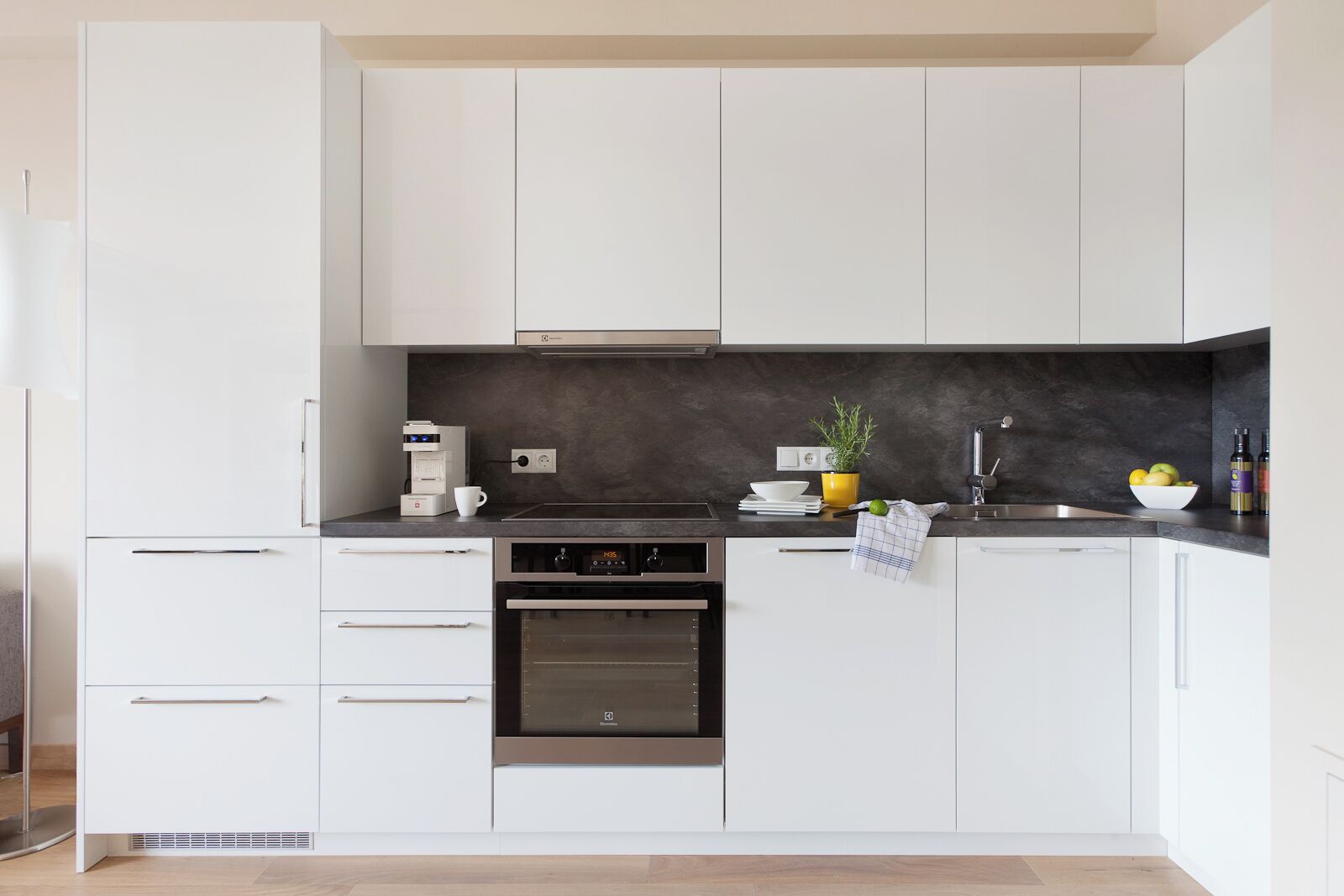A modern kitchen with white cabinets, a black countertop, a coffee machine, a plant, and a bowl of fruit.