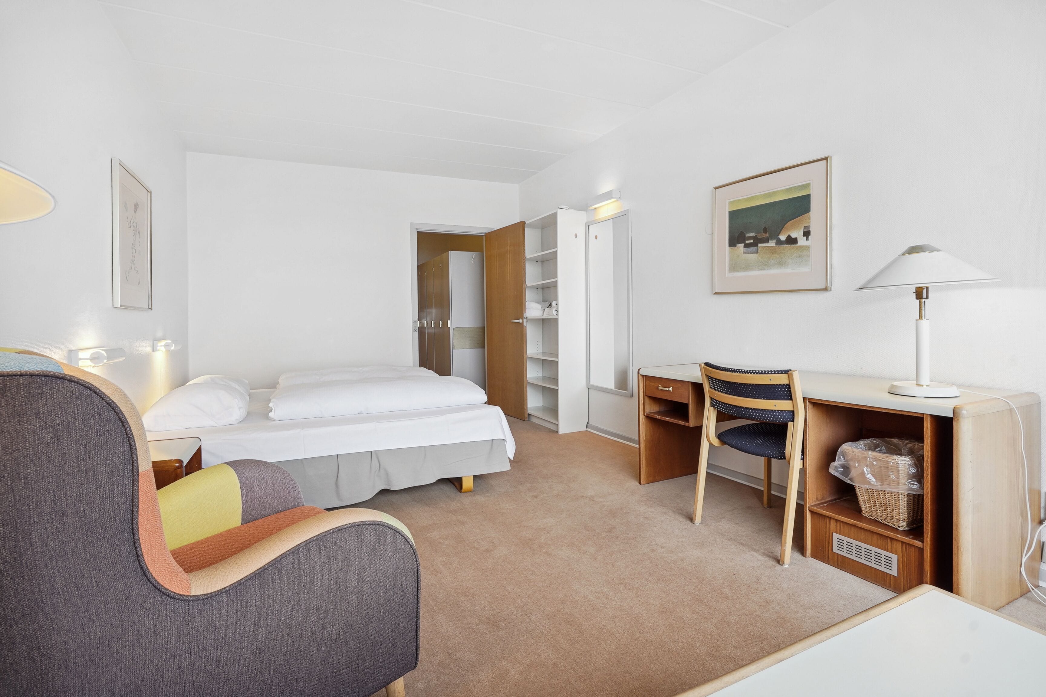 A hotel room with a bed, a desk, a chair, a wardrobe, and a lamp.