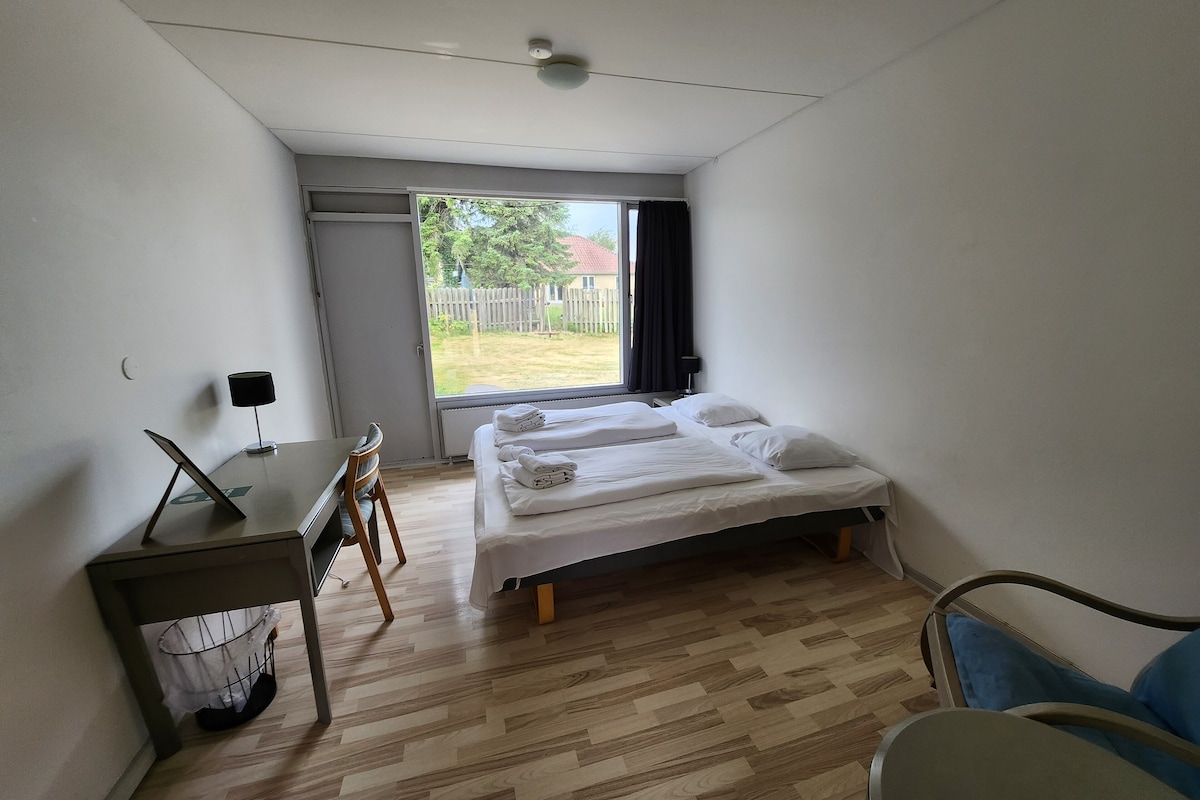 A bedroom with a bed, a desk, and a chair. There is a window with a view of a garden.