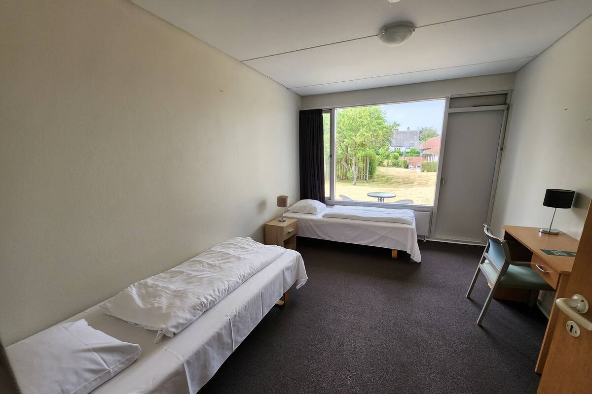 Habitación de hotel con dos camas individuales, un escritorio con silla, una mesita y una ventana con vistas al exterior.